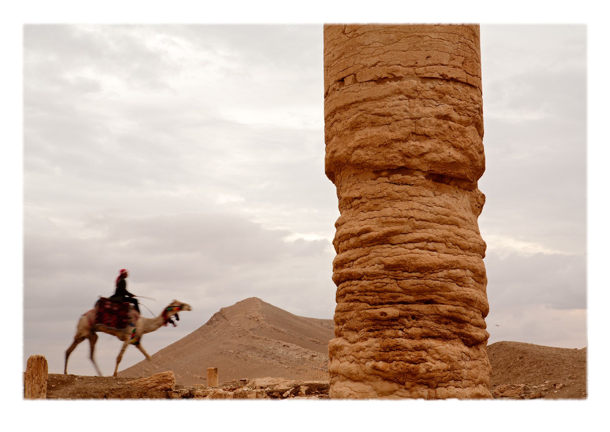 Dessicated windblown sandblasted and expressively eroded column behind which a man rides a colorfully bedecked camel. The tones and textures of erosion on once glorious specimens of  artisanal production are eloquent testaments to the relative nature of evanescence.