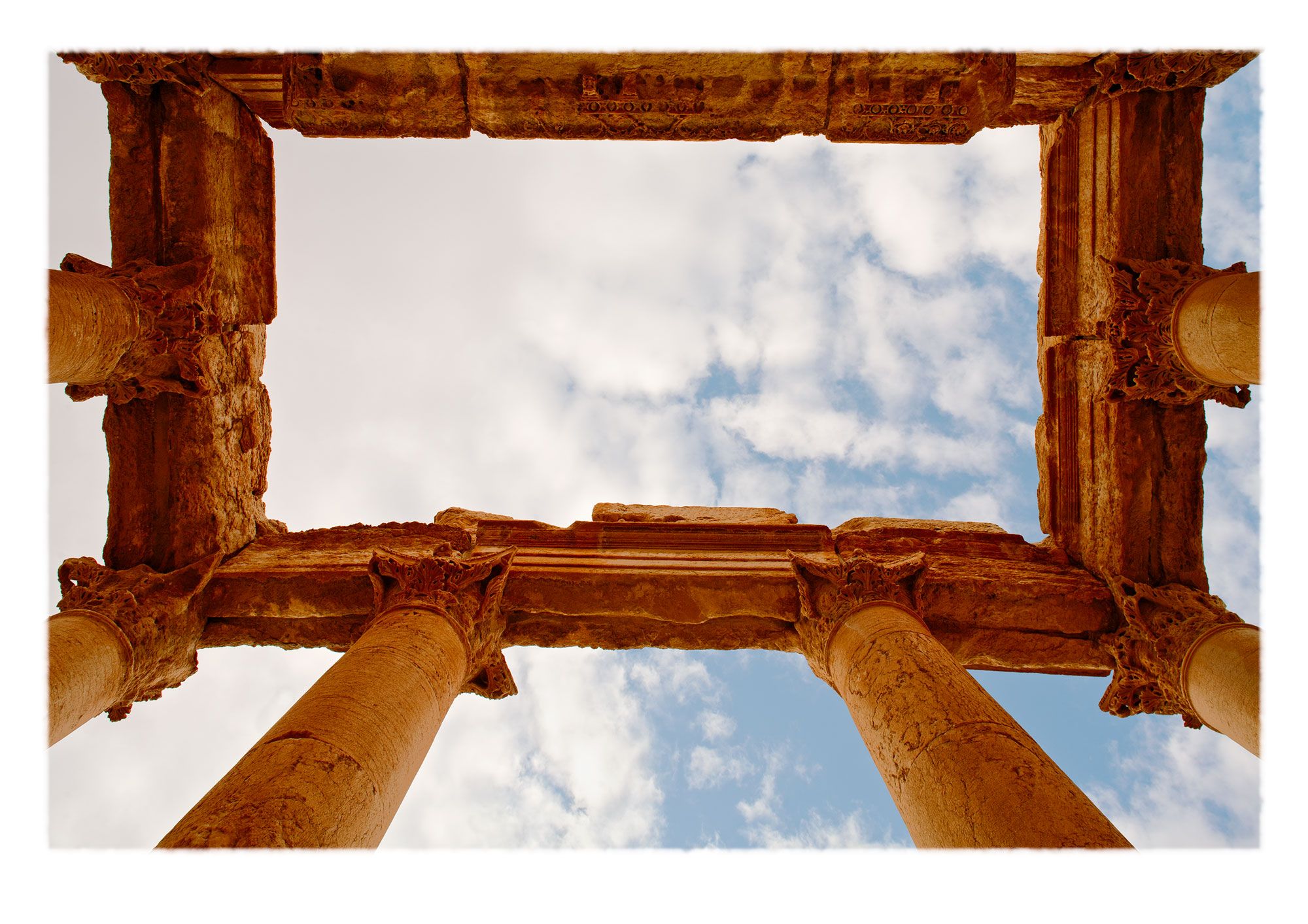 View upwards through Corinthian capitals of furled acanthus leaves with eroded entablature, sky and clouds. Destroyed Palmyra, Syria.